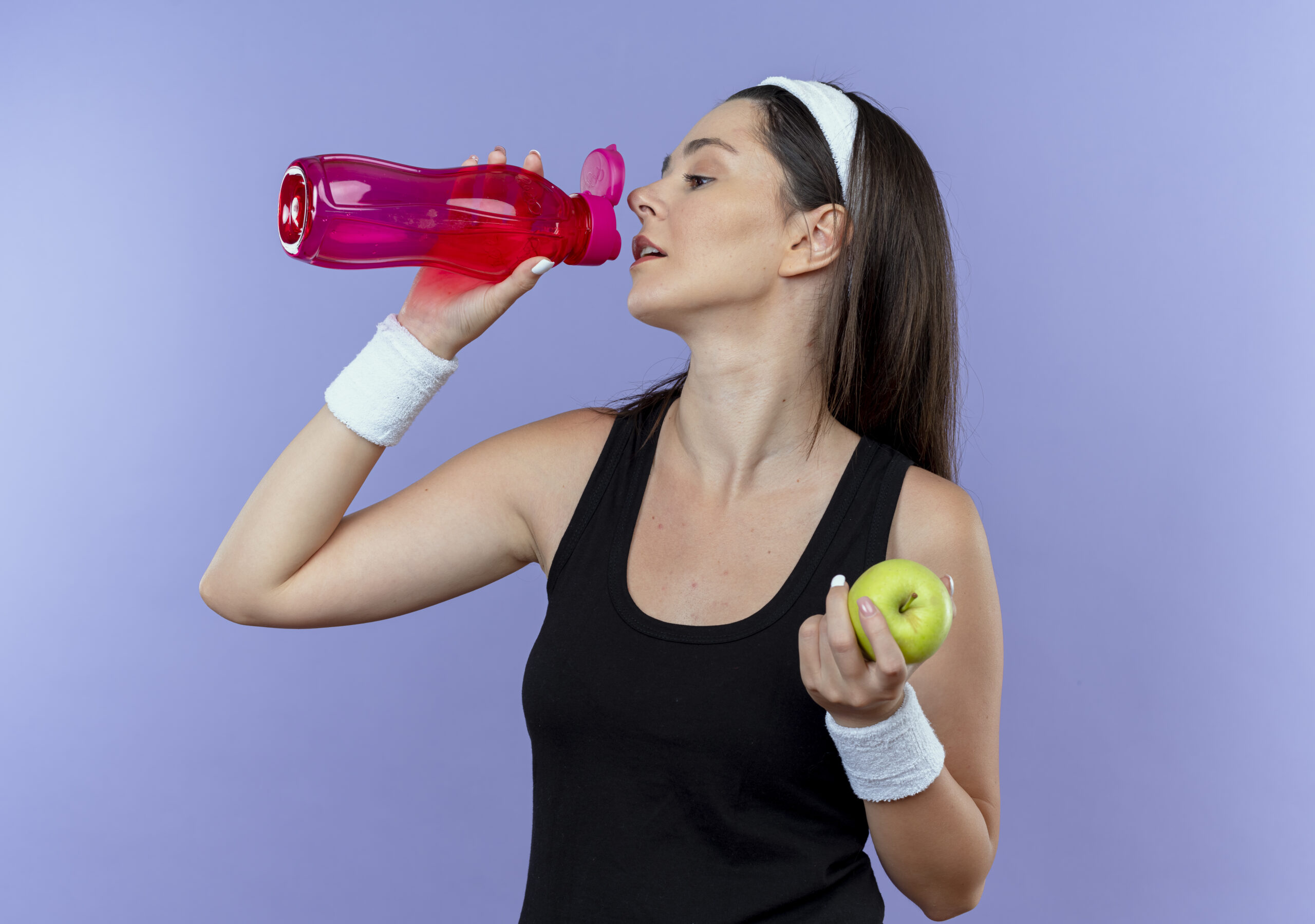 young-fitness-woman-headband-holding-green-apple-drinking-water-after-workout-standing-blue-wall