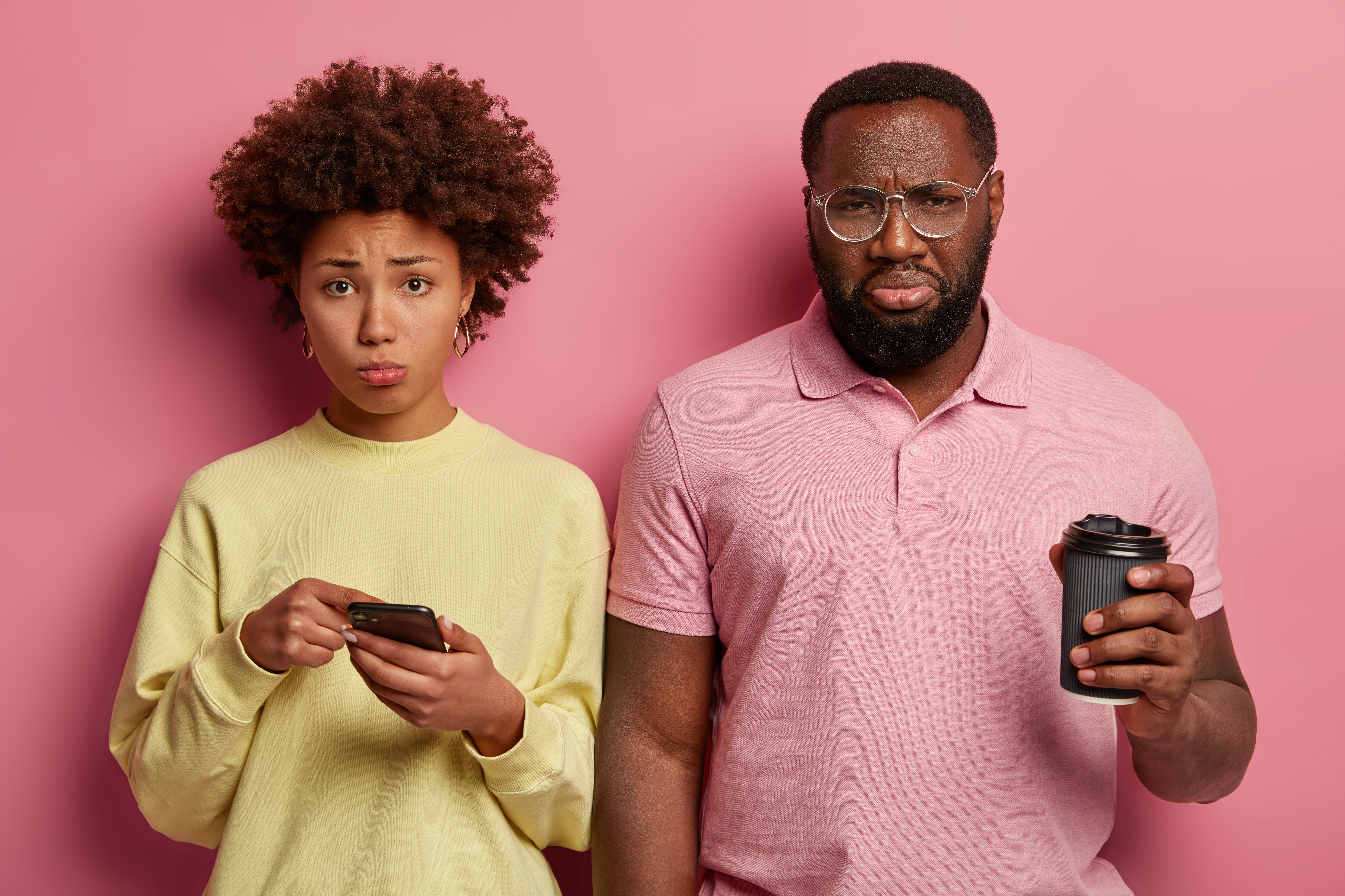 Photo of desperate sullen Afro American couple read bad news in social networks, drink takeaway coffee, sad woman points at display of smartphone, has some problems, isolated over pastel pink wall