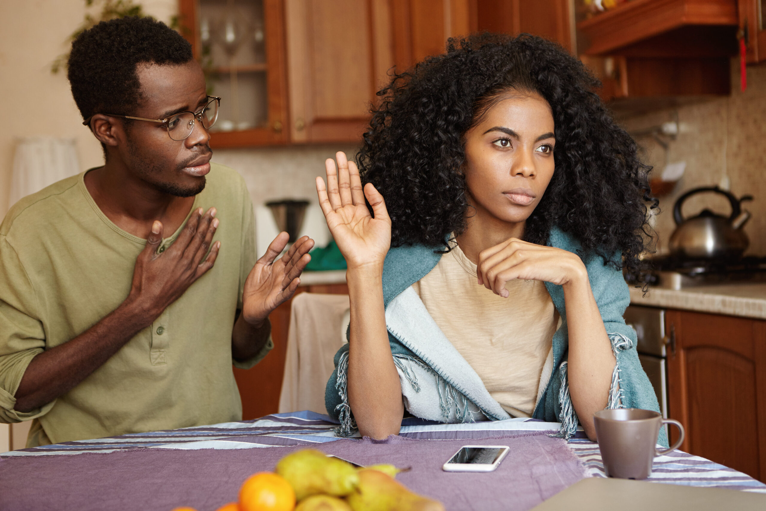 Sad African-American male in glasses holding hand on chest, apologizing to his angry and mad wife, who ignoring him, refusing to forgive him for cheating on her. People and relationships concept