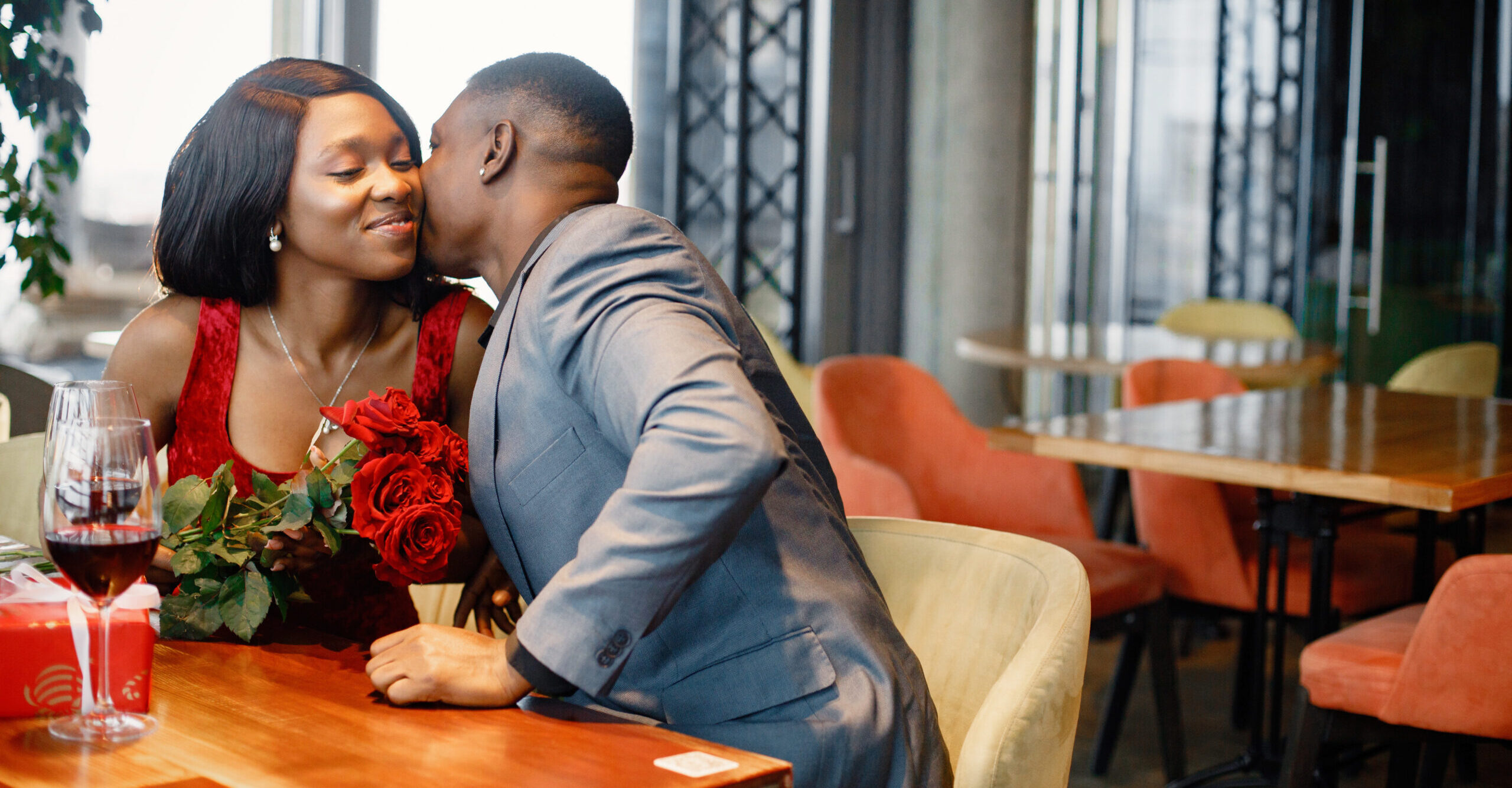 Couple enjoying day out at restaurant. Black man gifted a bouquet of red roses for a woman. Woman wearing red elegant dress and man blue costume.