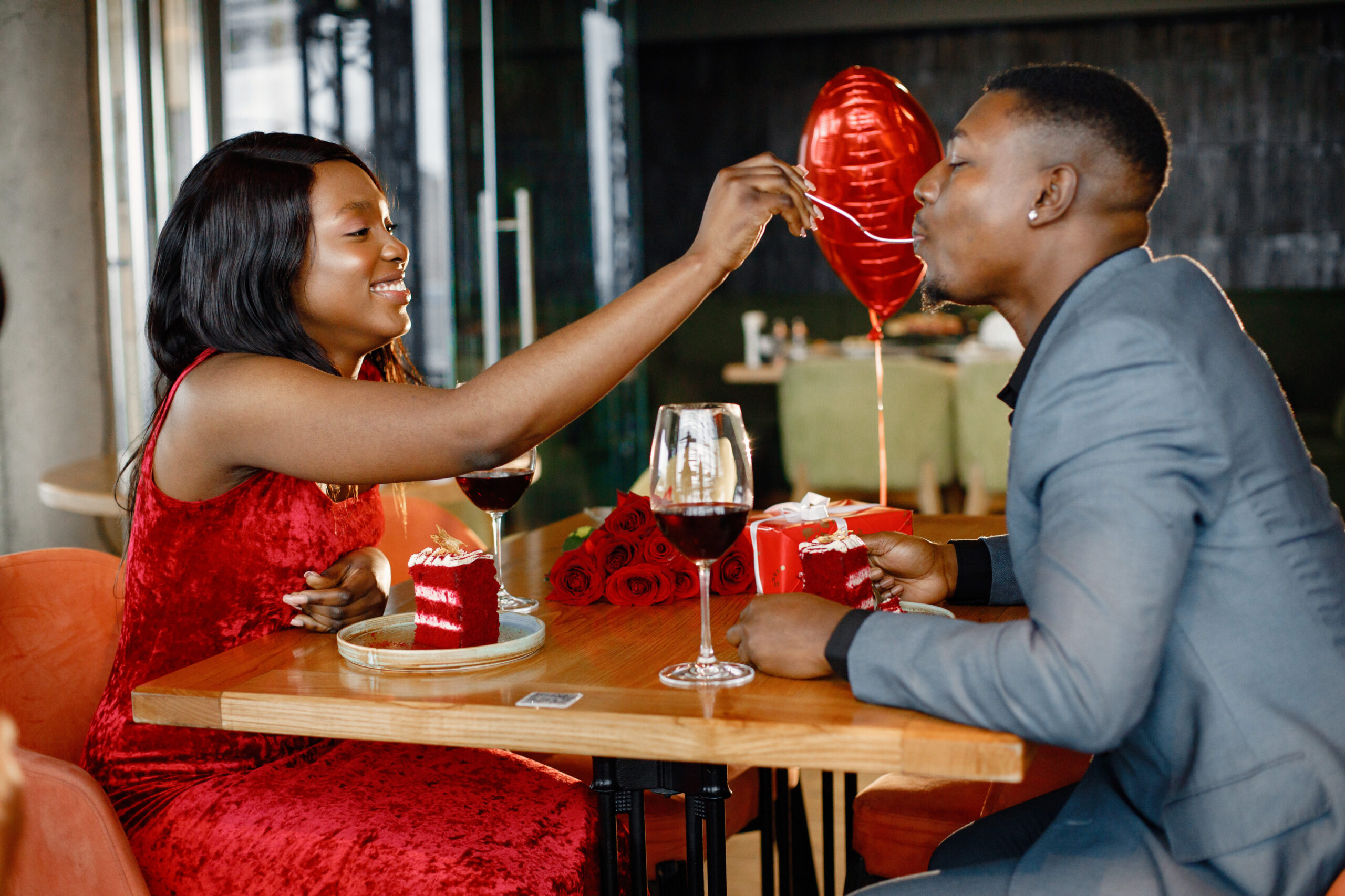 Couple enjoying day out at restaurant. Black man and woman drinking red wine and eating cakes. Woman wearing red elegant dress and man blue costume.
