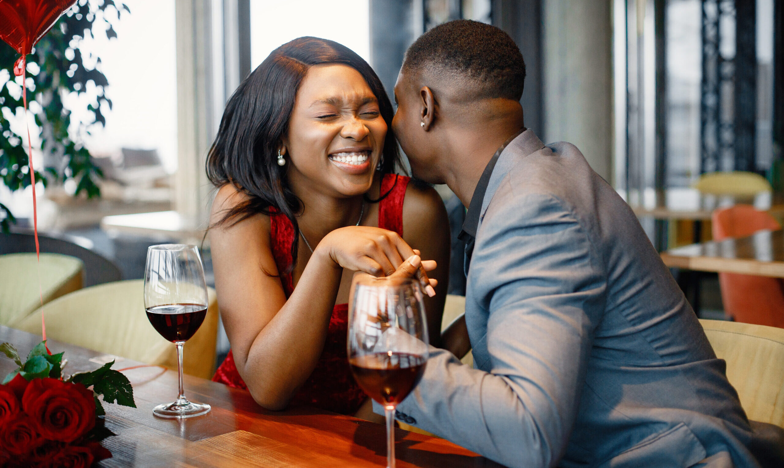 Couple enjoying day out at restaurant. Black man and woman talking and holding hands. Woman wearing red elegant dress and man blue costume.